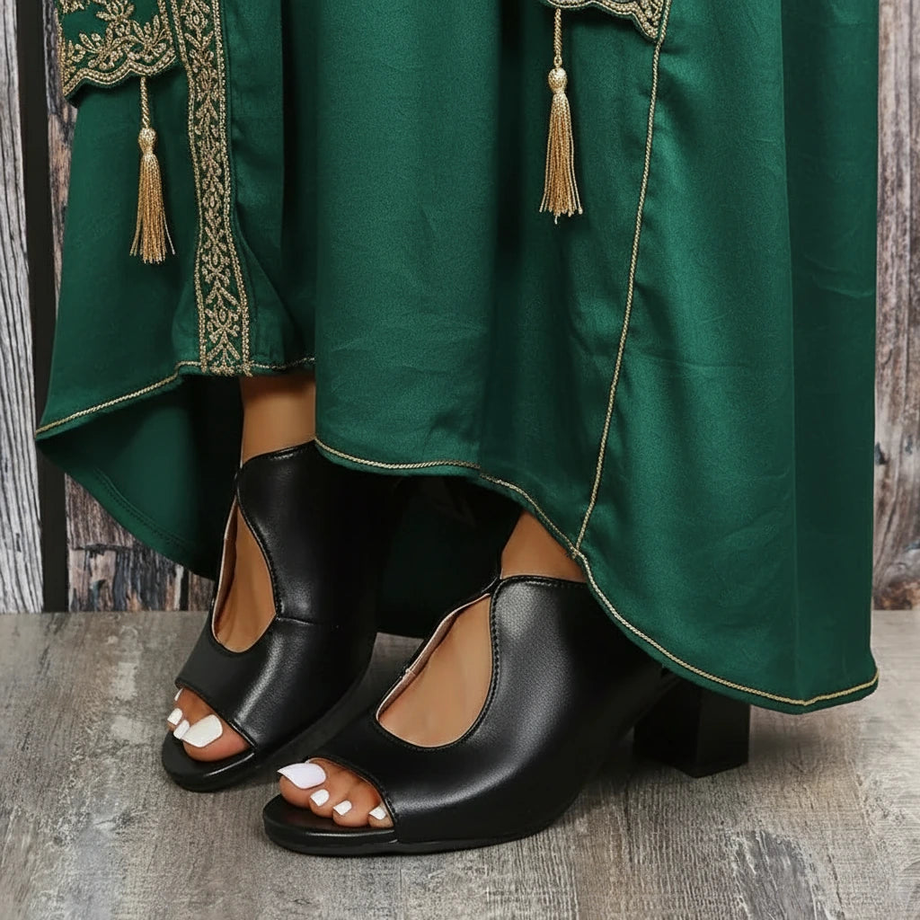 Black open-toe shoes worn with a green traditional outfit on a wooden floor.